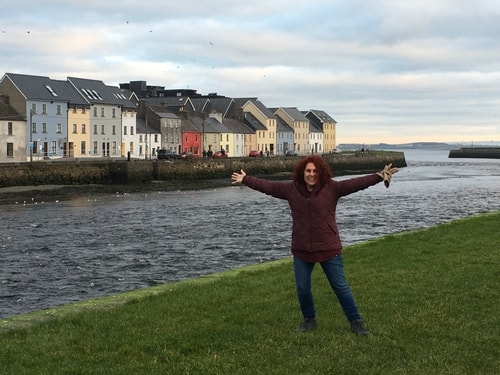 author standing with arms out wide in front of river, colourful buildings on the other side, in Galway