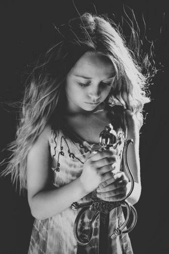 black and white image of young girl with long flowing hair looking down at the sword in her hands.